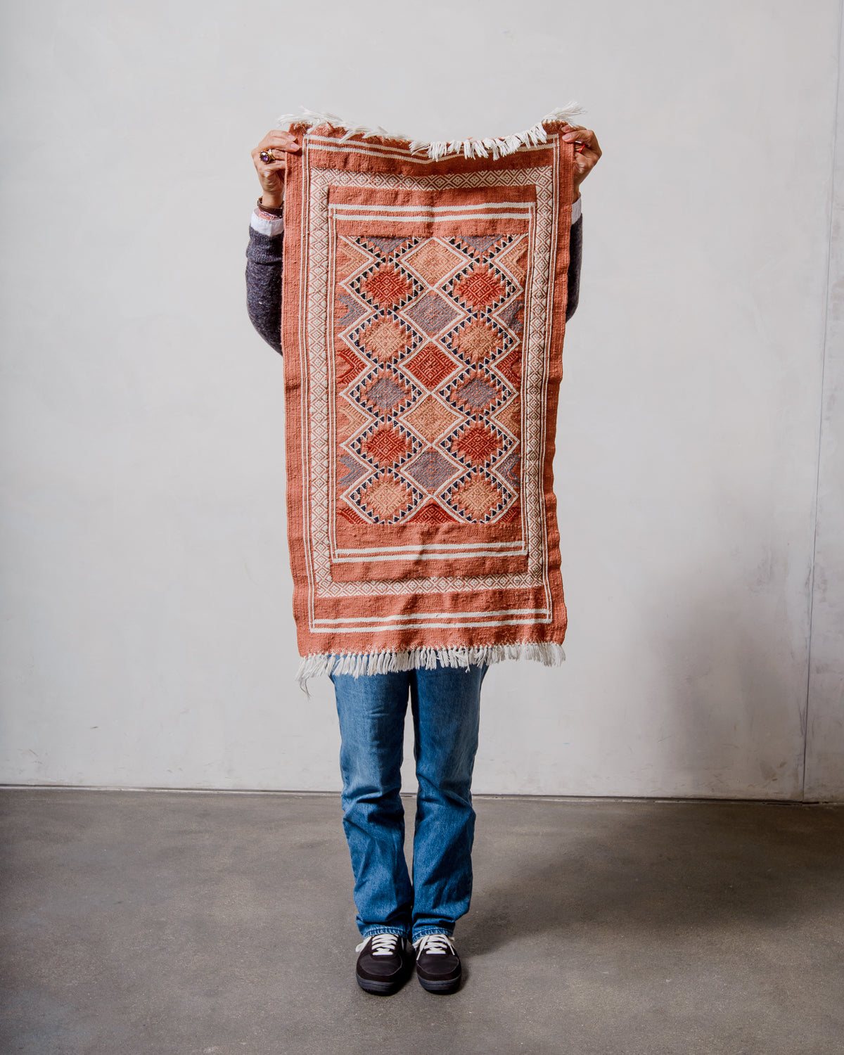 Small Berberlin Vintage Kelim Rug in shades of terracotta and white being held up in front of a white wall. 
