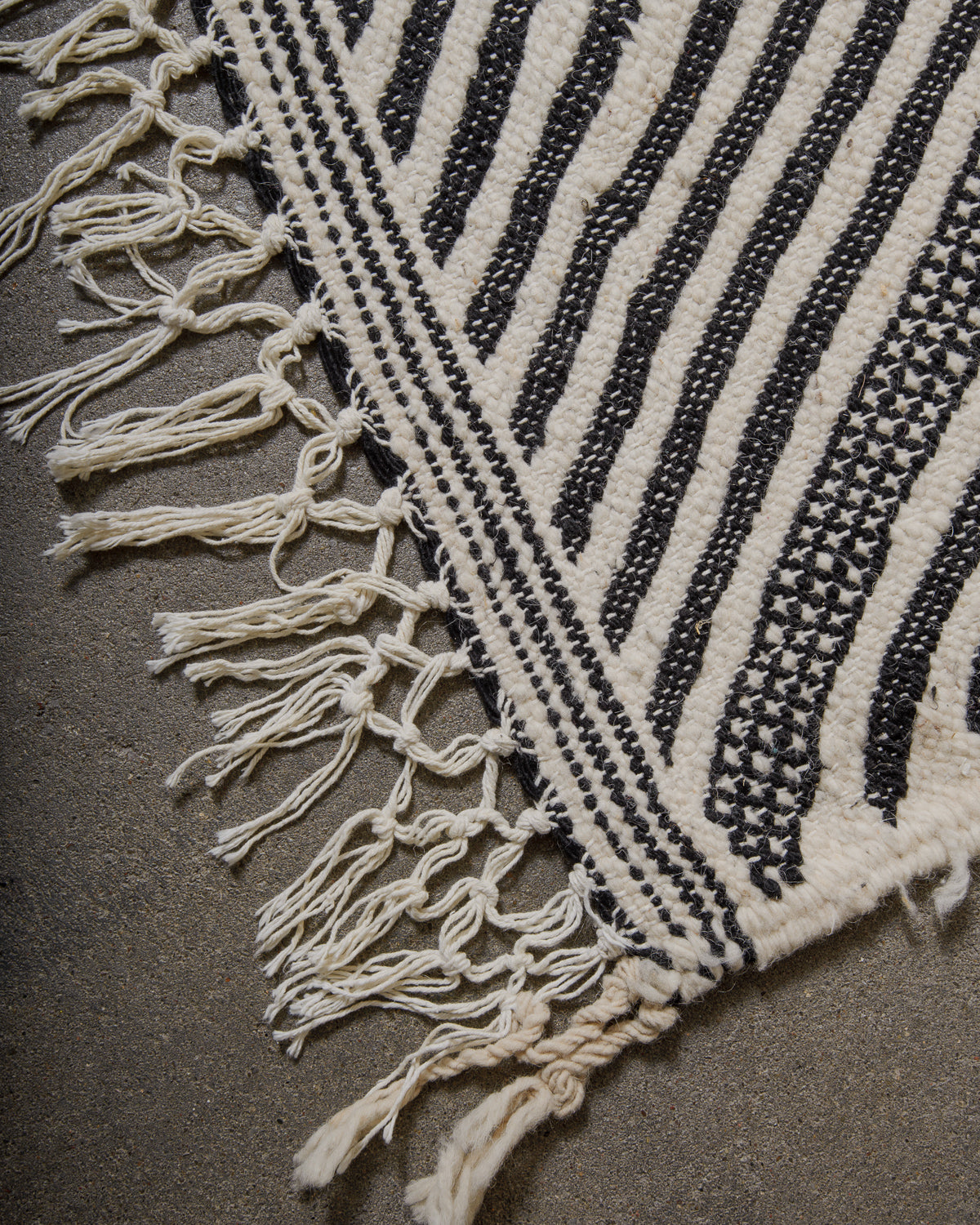 Close-up of a black and white Kelim rug with tassels on a dark surface.
