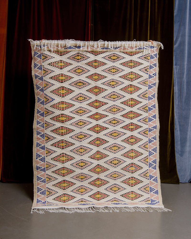 Kelim rug with diamond pattern held up in front of velvet curtains in different colours.