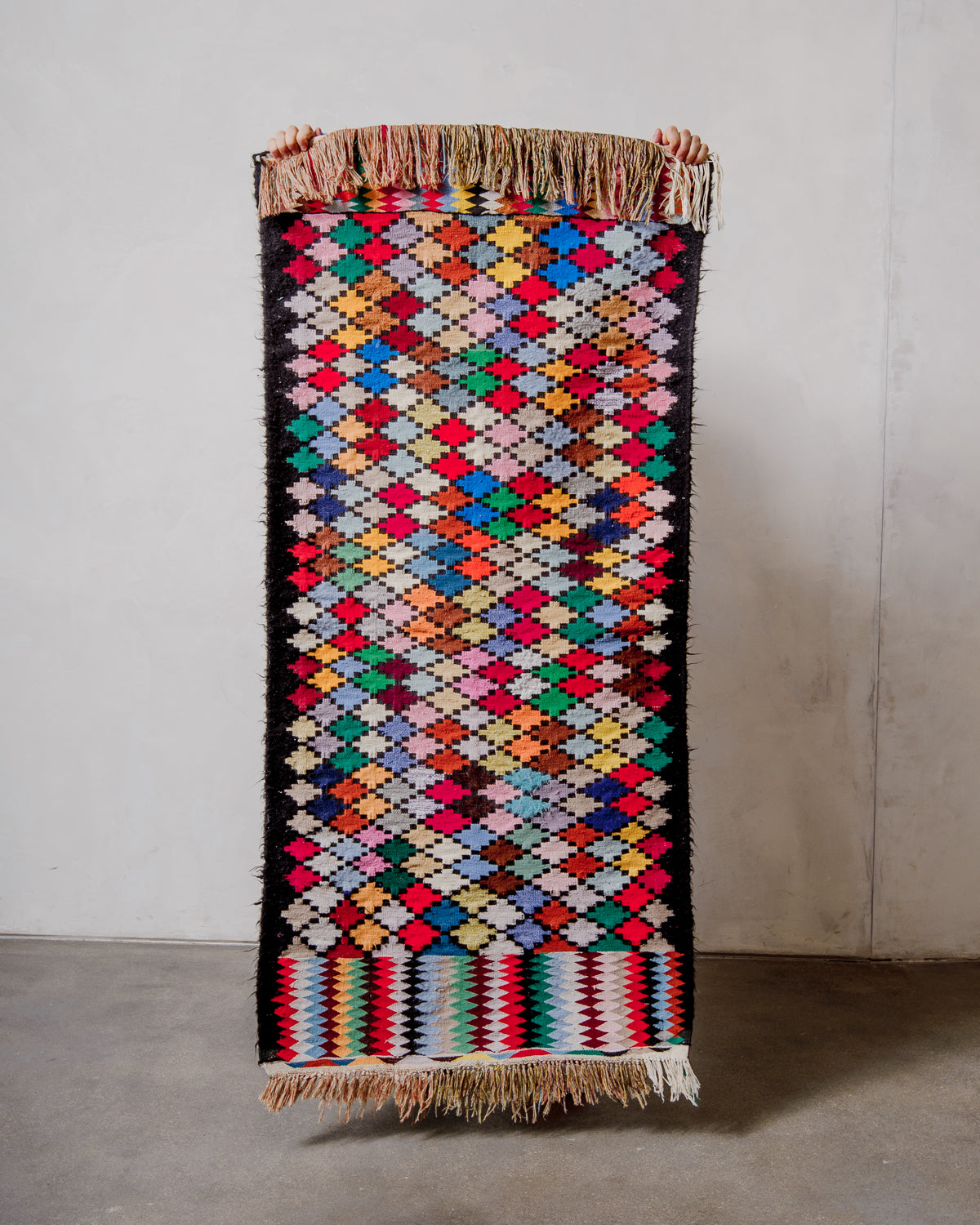 Vintage Kelim runner rug with a brightly coloured harlequin design and black borders, being held up in front of a white wall.