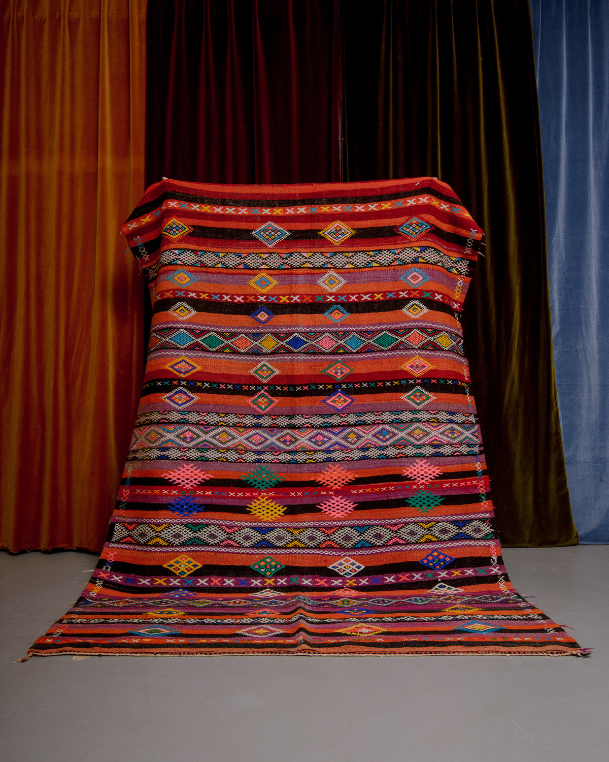 Vintage Kelim rug in bright orange, red, green and pink being held in front of velvet curtains. 