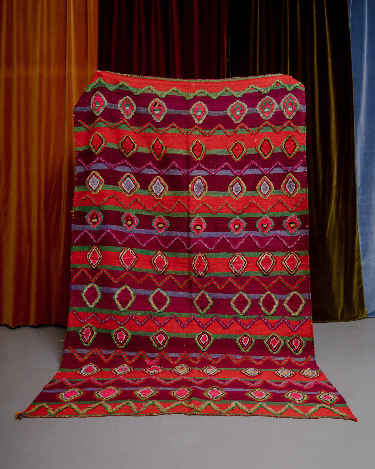Vintage Kelim rug in bright red, green and burgundy being held in front of velvet curtains. 