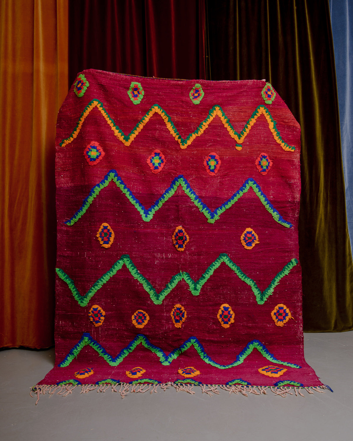 Vintage Kelim rug in bright red, green, orange and blue being held in front of velvet curtains. 