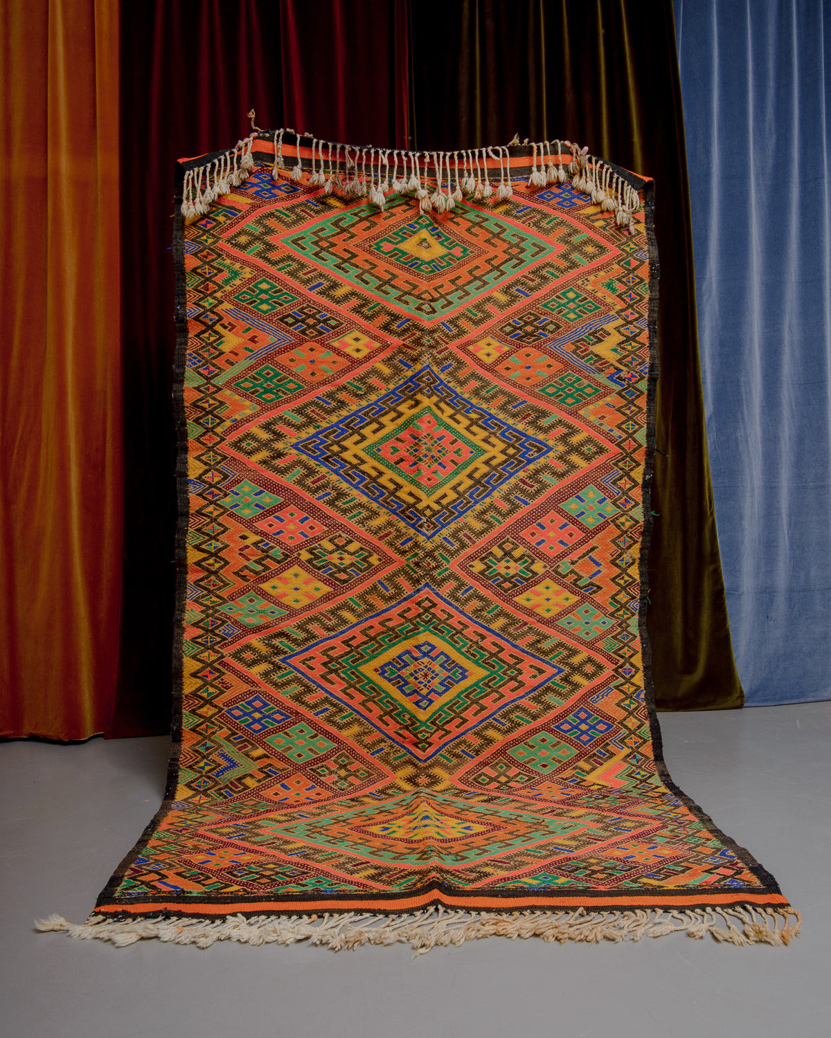 Vintage Kelim rug in bright yellow, orange, blue and green held in front of velvet curtains. 