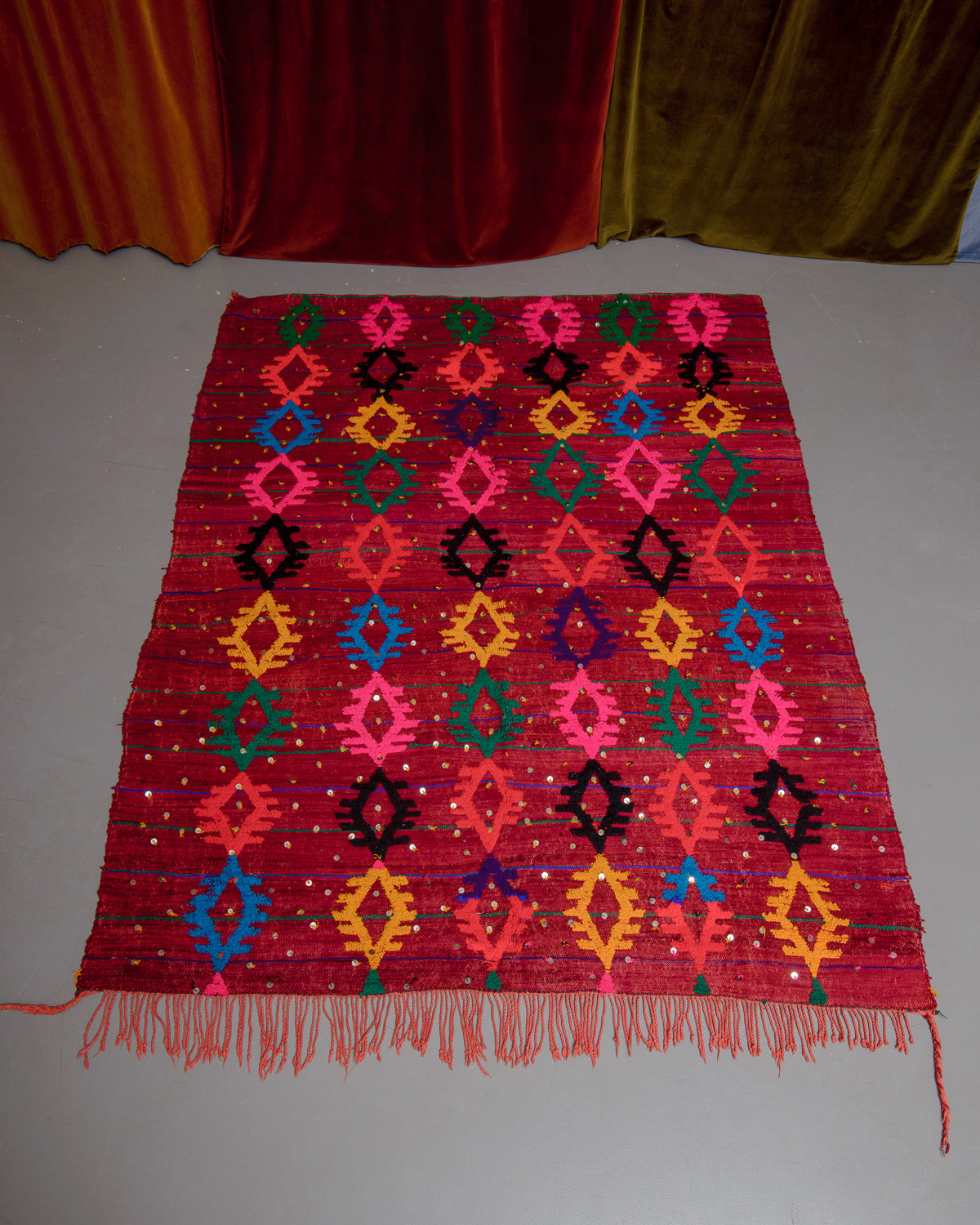 Vintage Kelim rug in bright red, green, orange, pink and blue on grey floor in front of velvet curtains.