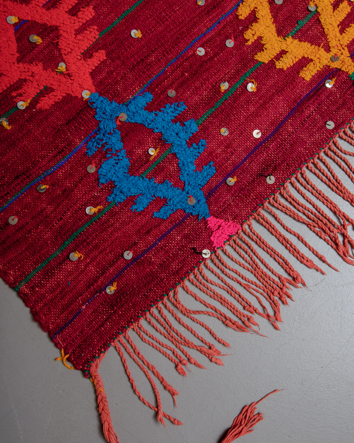 Corner of vintage kelim rug in red, blue, yellow and orange with red tassels on grey floor. 