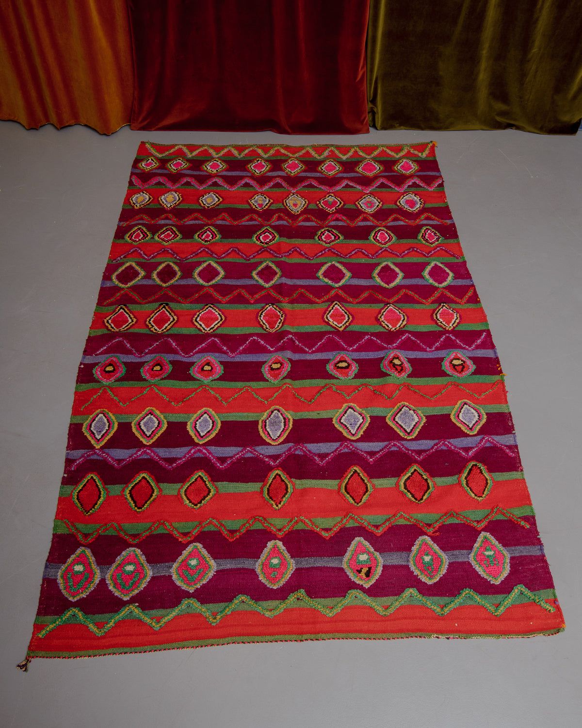 Vintage Kelim rug in bright red, green and burgundy laying on grey floor in front of velvet curtains. 