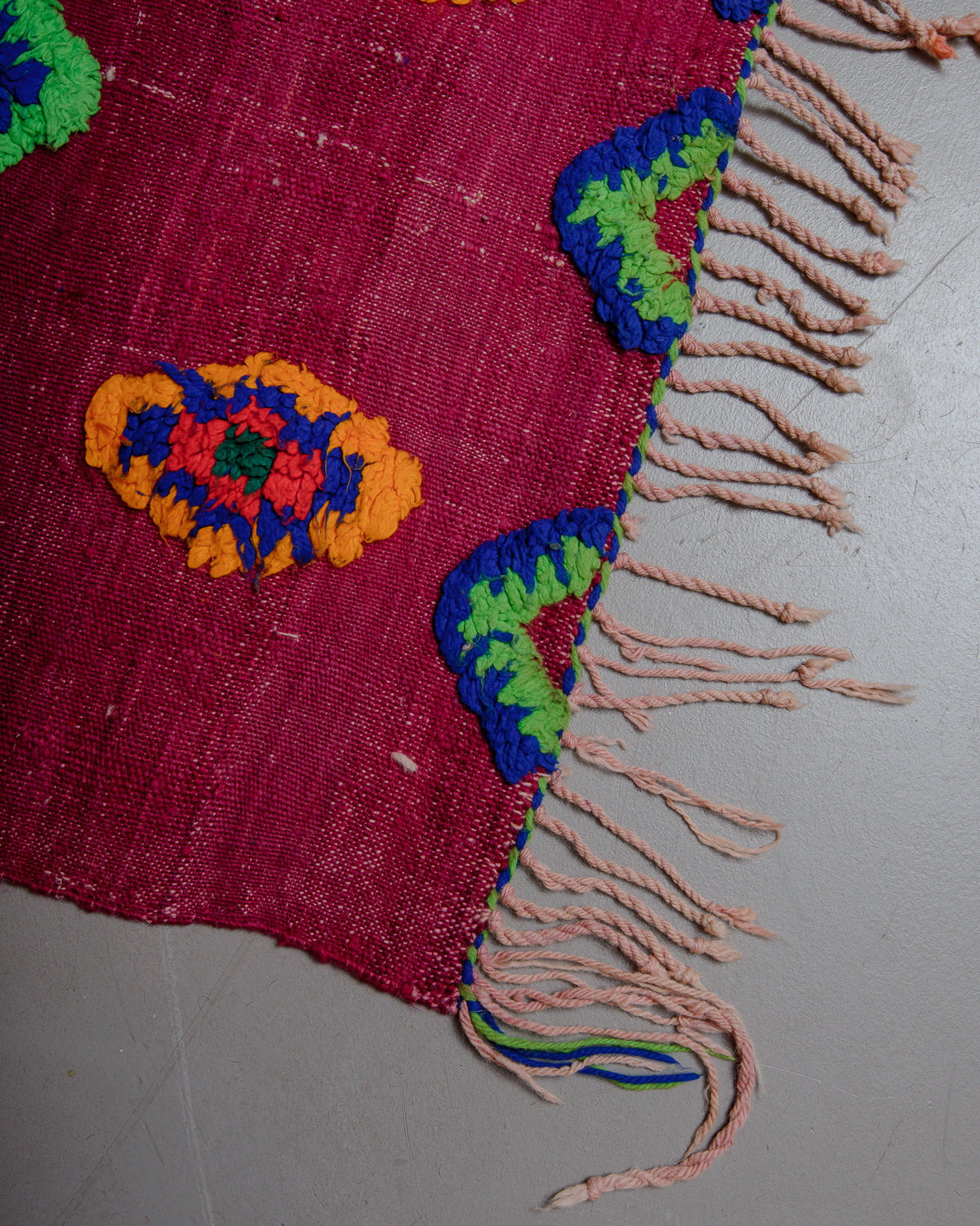 Corner of vintage kelim rug in red, green, blue and orange with tassels on grey floor. 