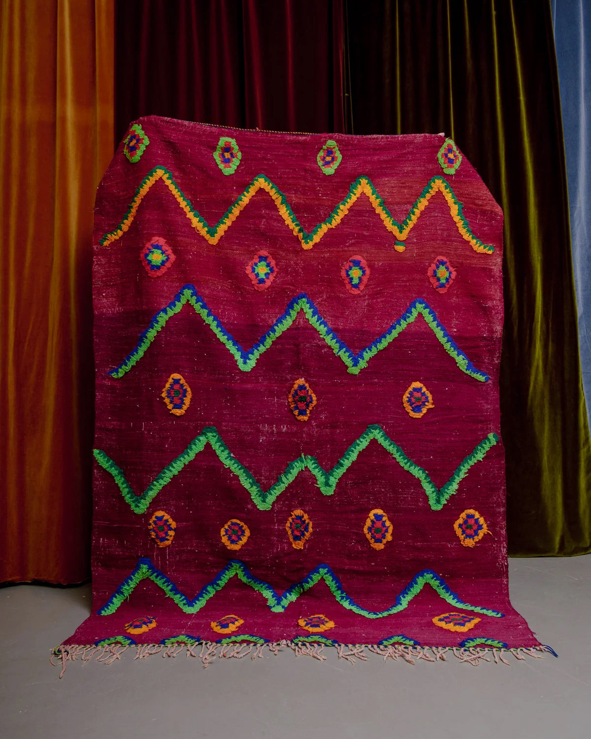 Vintage Kelim rug in bright red, green, orange and blue being held in front of velvet curtains. 