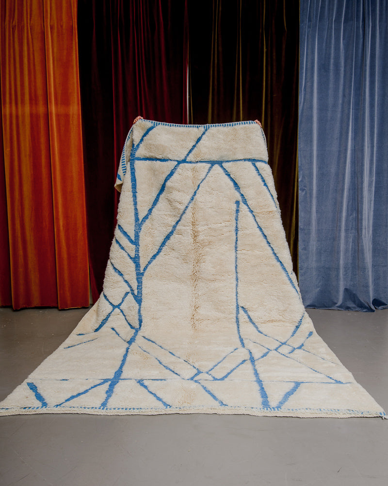 Cream Berberlin M'rirt with bright teal line design being held up in front of orange, red and blue velvet curtains.
