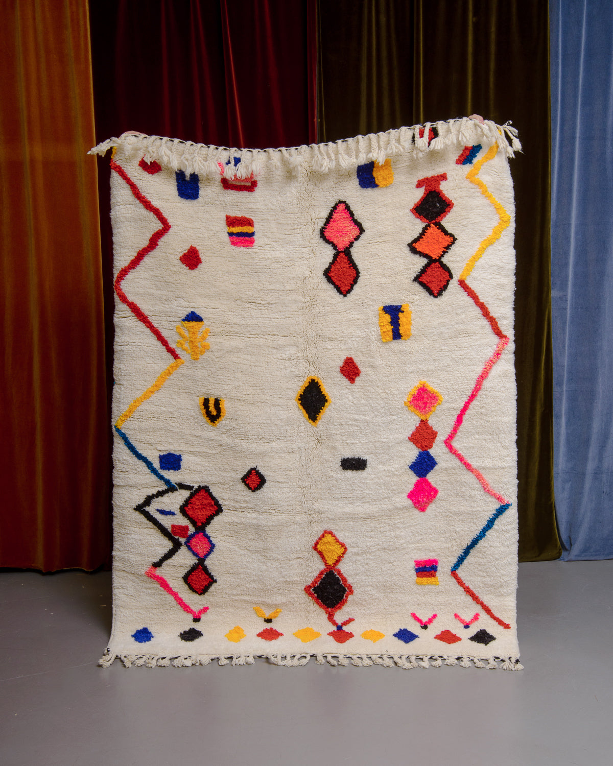 Berberlin Finest rug with a thick cream pile and colourful design in cotton being held in front of velvet curtains. 