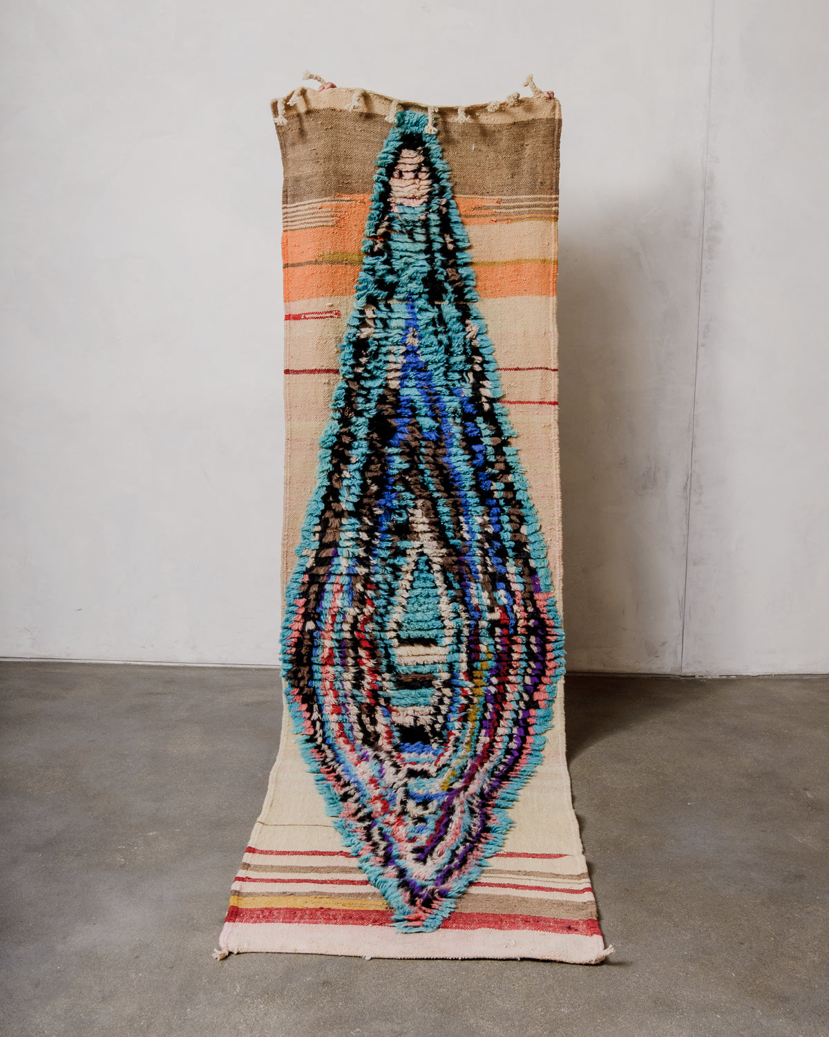 Berberlin vintage runner rug made of brightly coloured wool, being held up in front of a white wall and concrete floor. 
