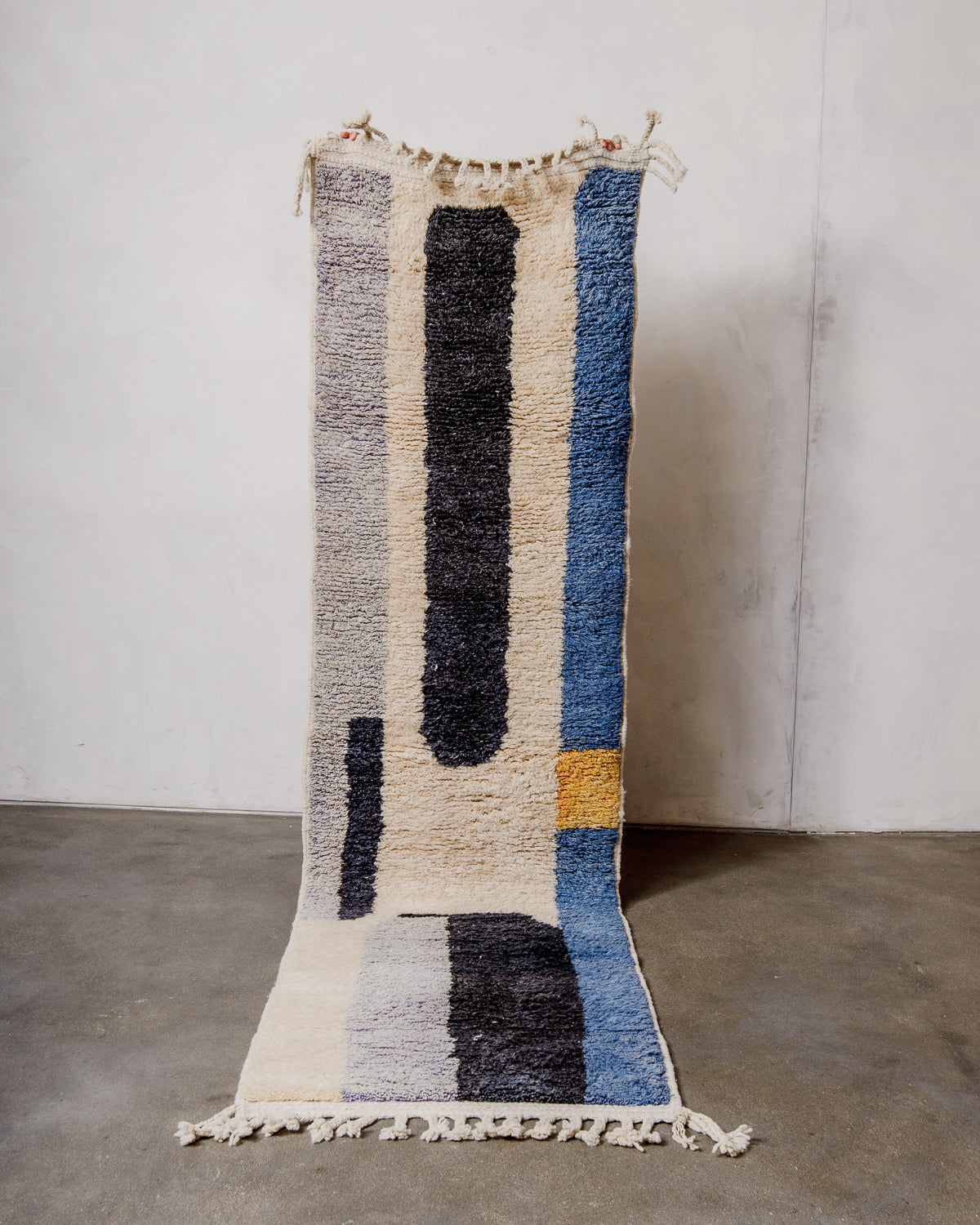 Berberlin runner rug made of sheep's wool, being held up in front of a white wall and concrete floor. 