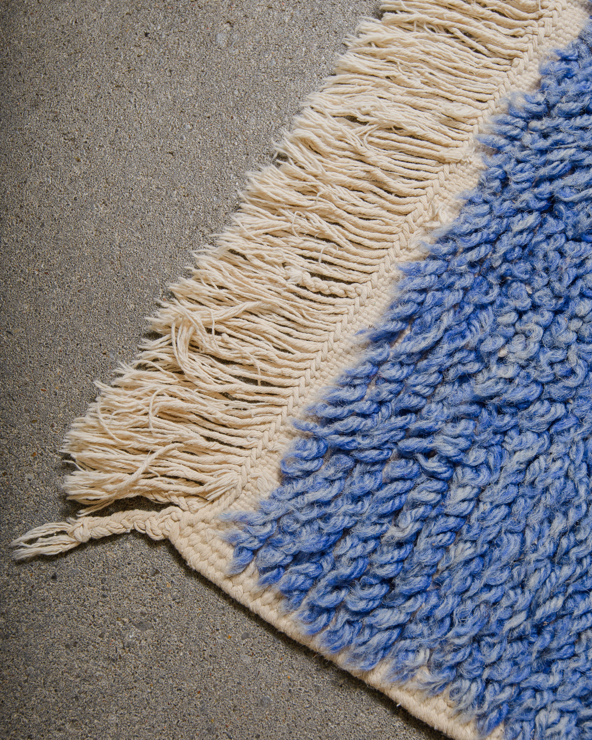Close-up of a blue and cream  textured rug corner with tassels on a concrete floor.