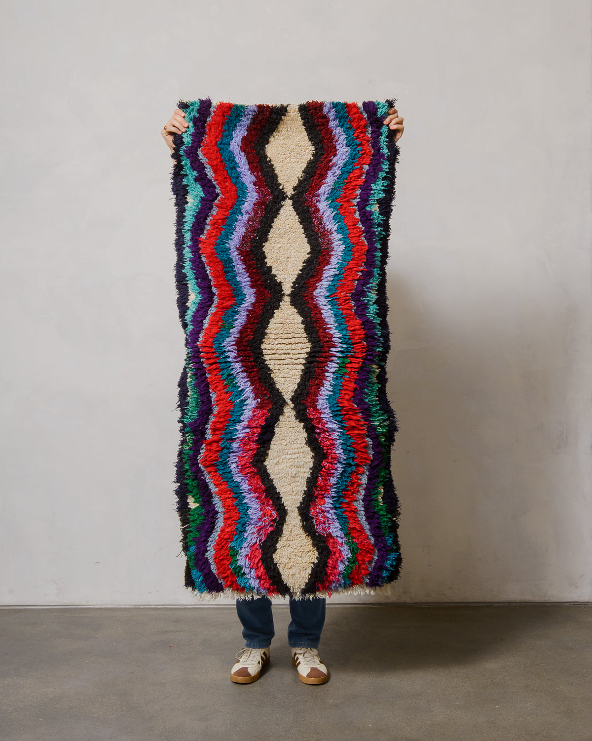 Person holding a colorful, patterned rug against a plain wall.