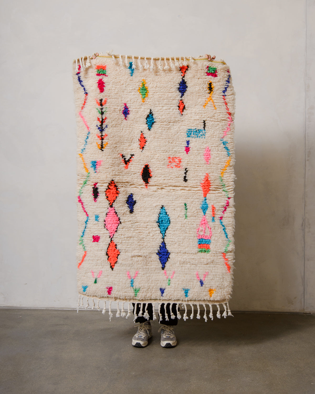 Berberlin Azilal rug in cream wool with colourful cotton  being held up in front of a white wall.