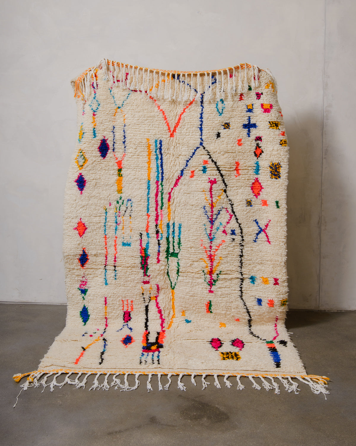 Berberlin Azilal rug made of fluffy cream wool and colourful design, held up in front of a white wall and cement floor.