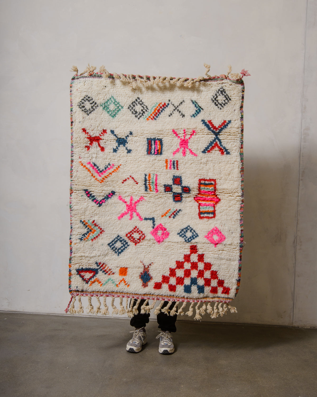 Small Berberlin Beniourain rug with a cream background and colourful design, being held up in front of a white wall. 