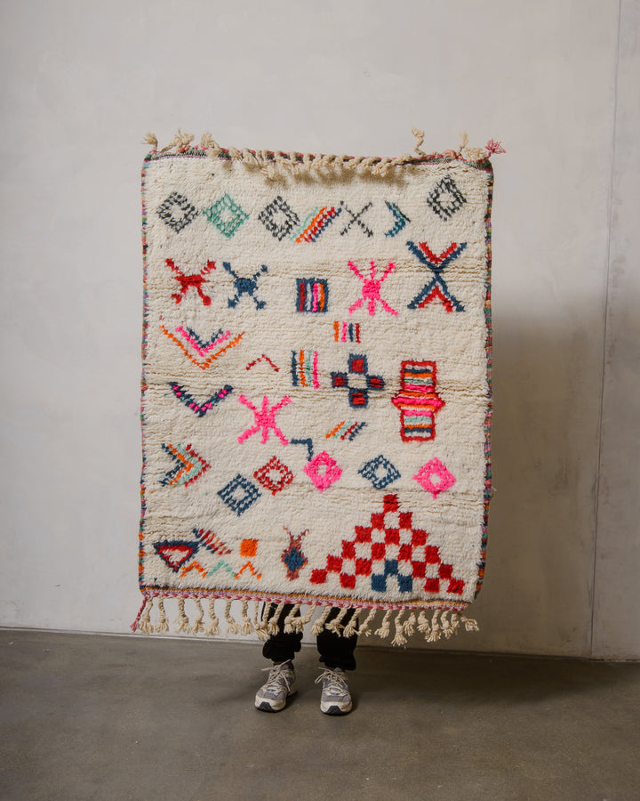 Small Berberlin Beniourain rug with a cream background and colourful design, being held up in front of a white wall. 