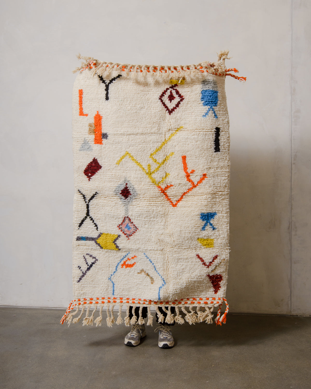 Small Berberlin Beniourain rug with a cream background and colourful design, being held up in front of a white wall. 