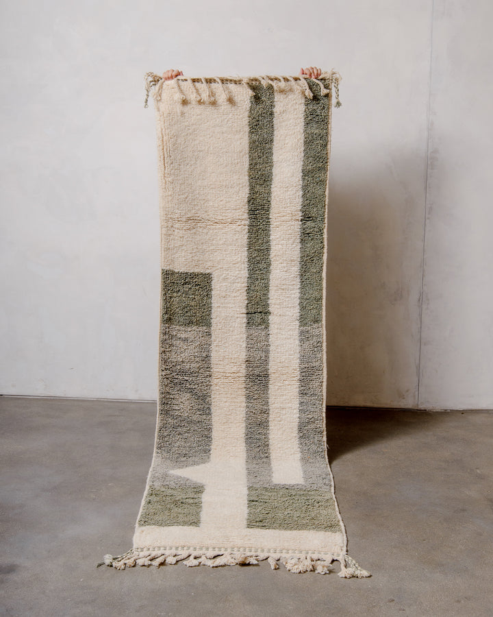 Berberlin Berber runner rug in cream and sage green wool, being held up in front of a white wall. 