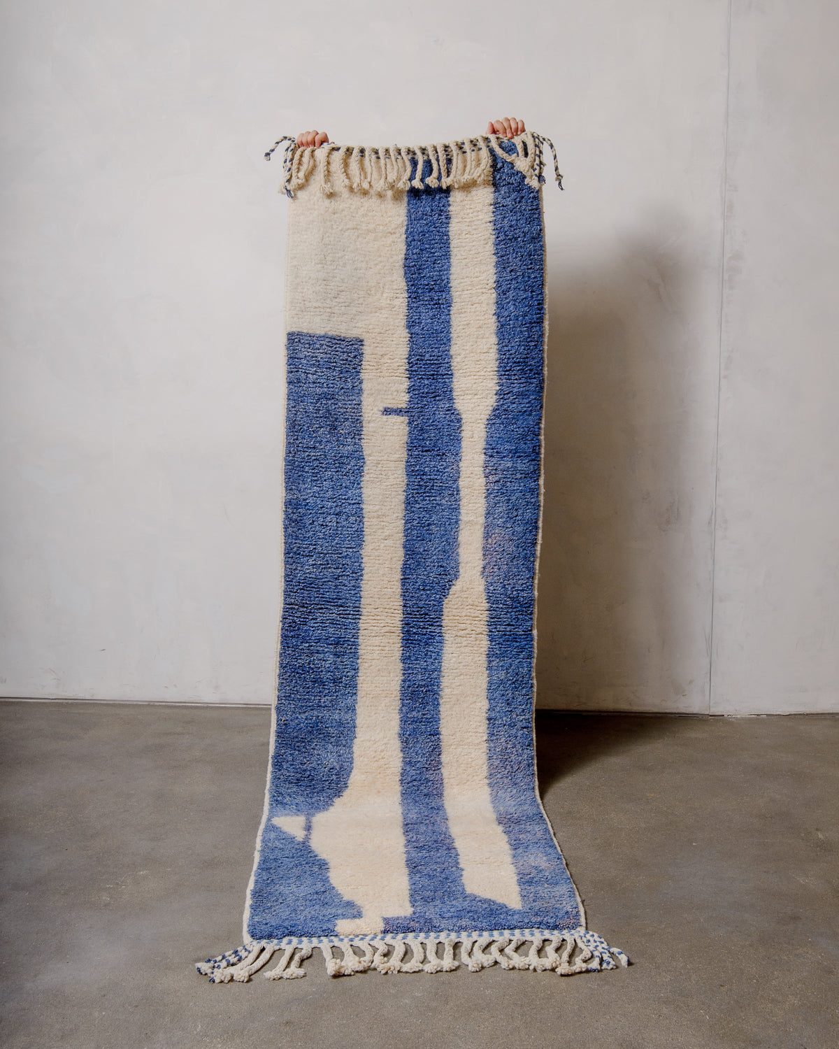 Berber runner rug made of blue and cream sheep's wool being held up in front of a white wall.