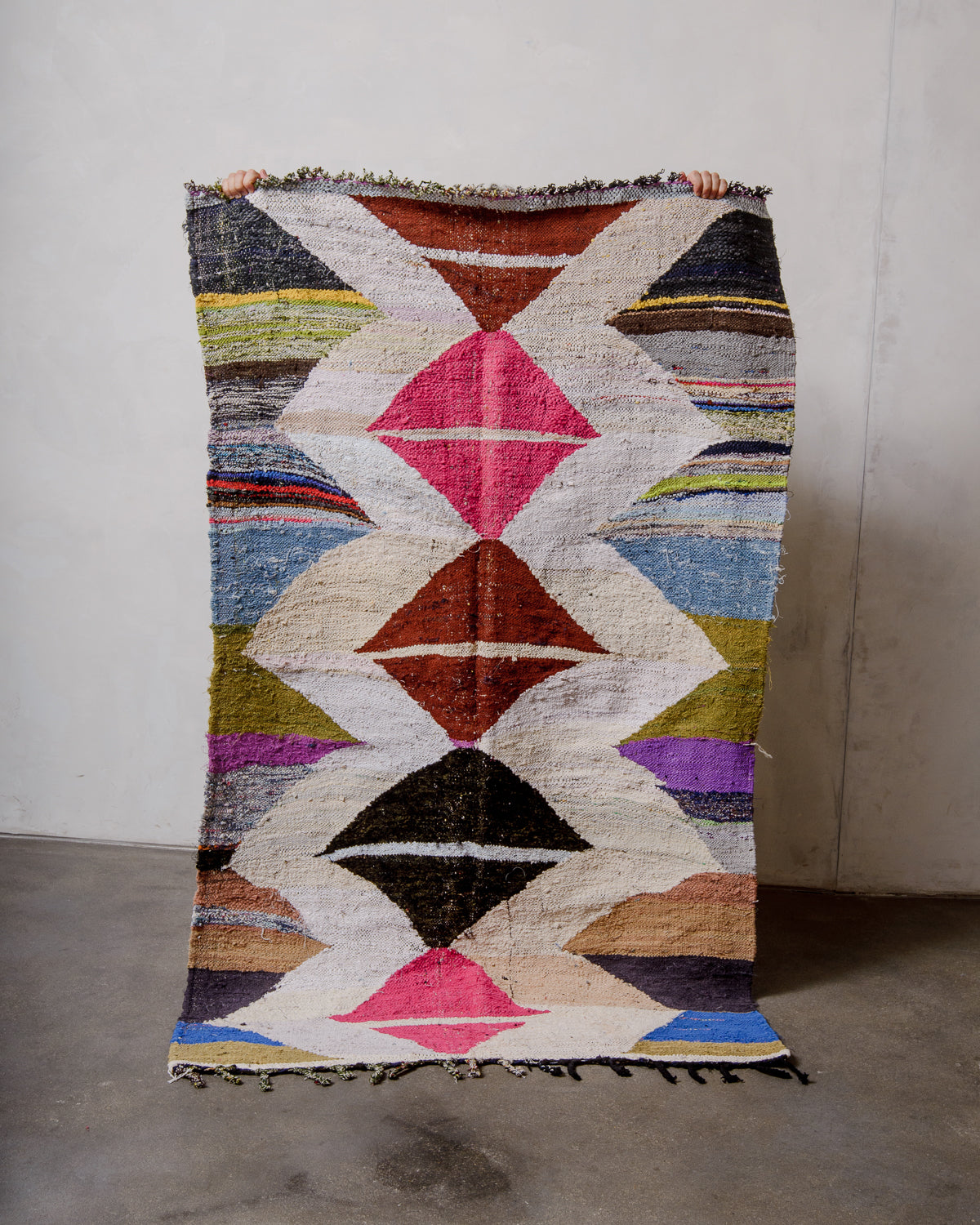Berberlin vintage kelim rug made of recycled materials being held up by two hands in front of a white wall and concrete floor. 