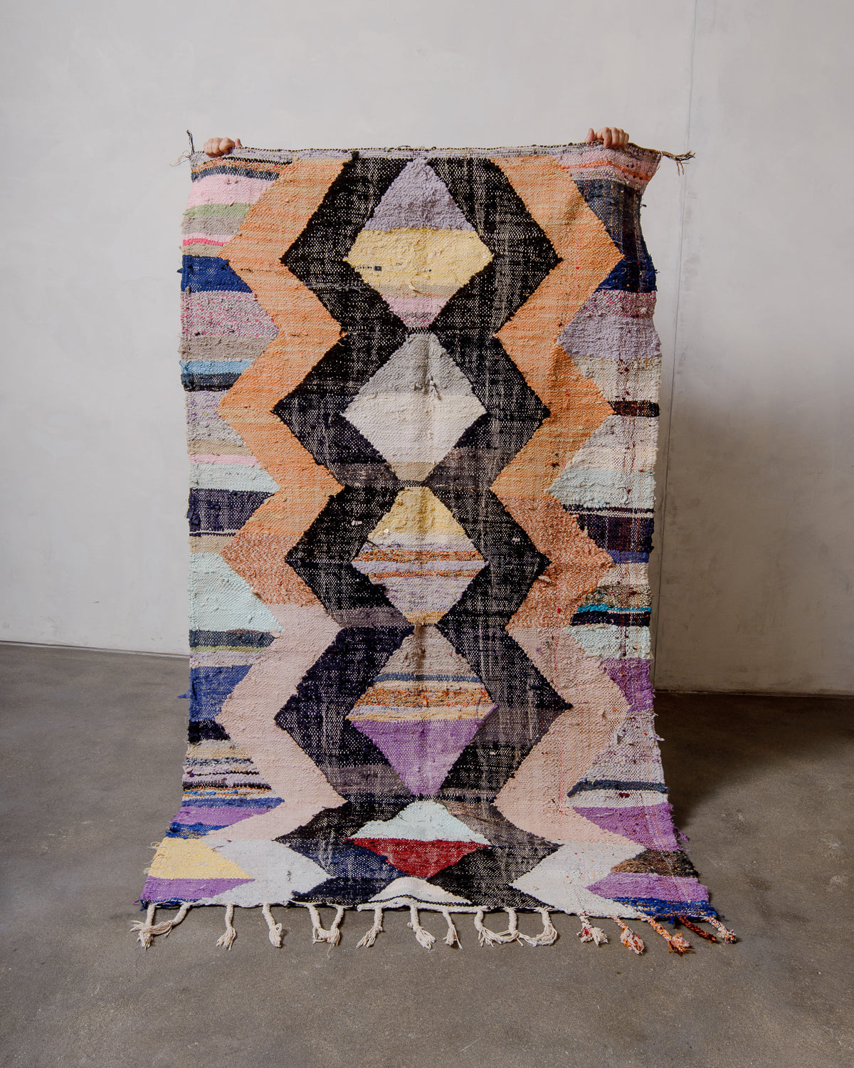 Berberlin vintage kelim rug made of recycled materials being held up by two hands in front of a white wall and concrete floor. 