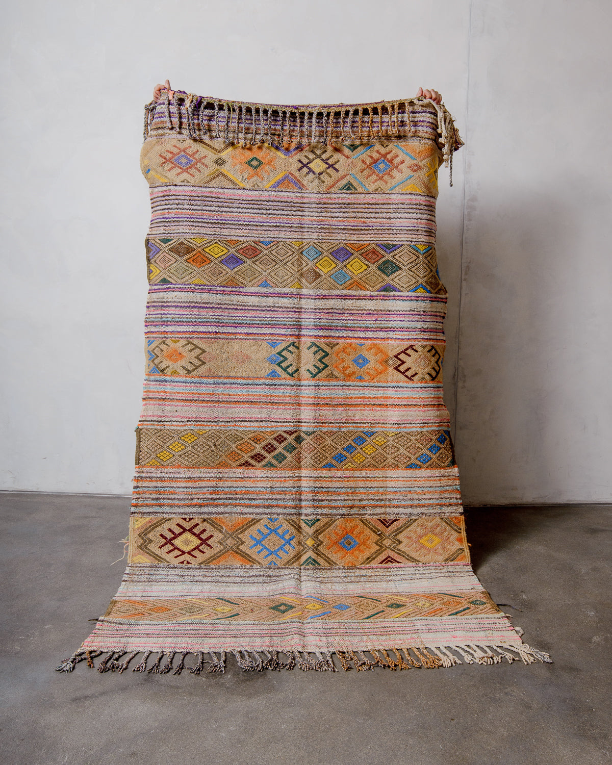 Berberlin vintage moroccan Kelim rug with colourful details being held up by two hands in front of a white wall and concrete floor. 
