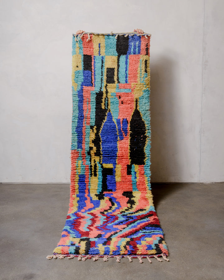 Berberlin vintage runner rug made of brightly coloured wool, being held up in front of a white wall and concrete floor. 