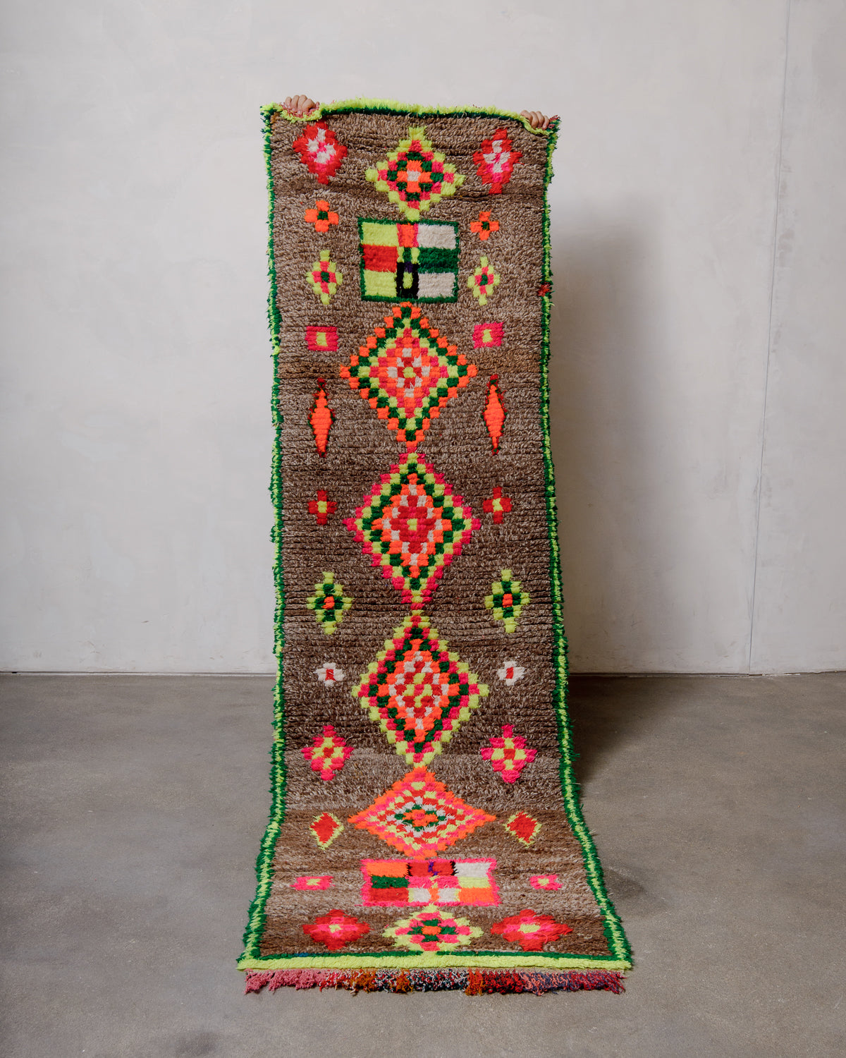 Berberlin vintage runner rug with brown wool and neon cotton, being held up in front of a white wall and grey concrete floor.