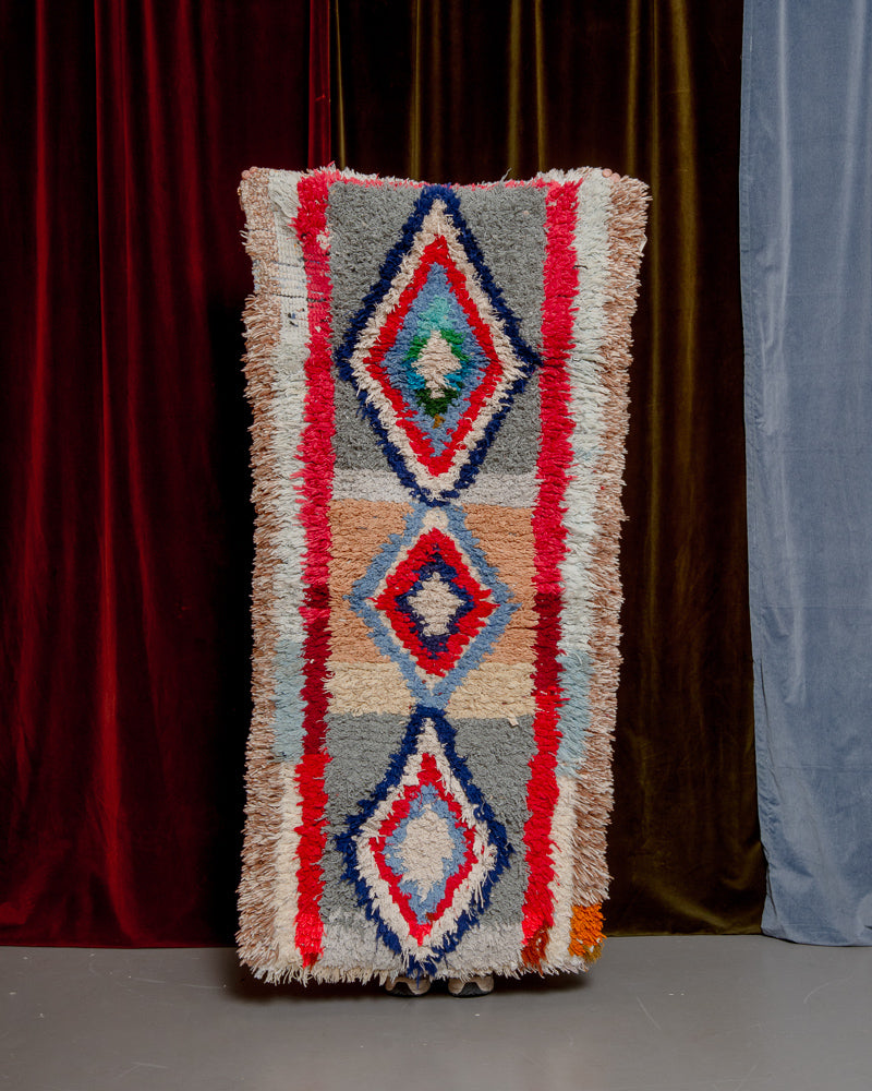 Vintage Berberlin boucherouite runner rug being held up in front of a velvet curtain. 