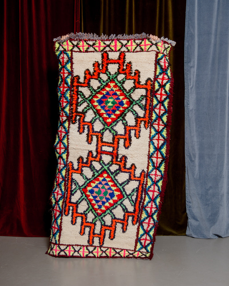 Vintage Berberlin boucherouite runner rug being held up in front of a velvet curtain. 