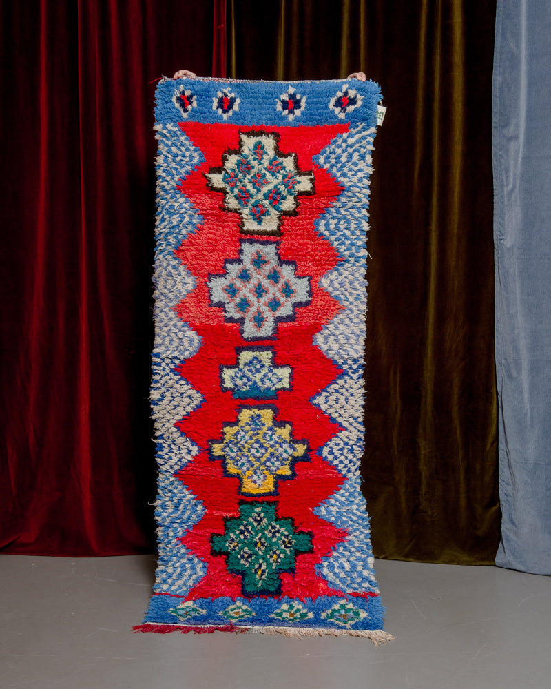 Vintage Berberlin boucherouite runner rug being held up in front of a velvet curtain. 