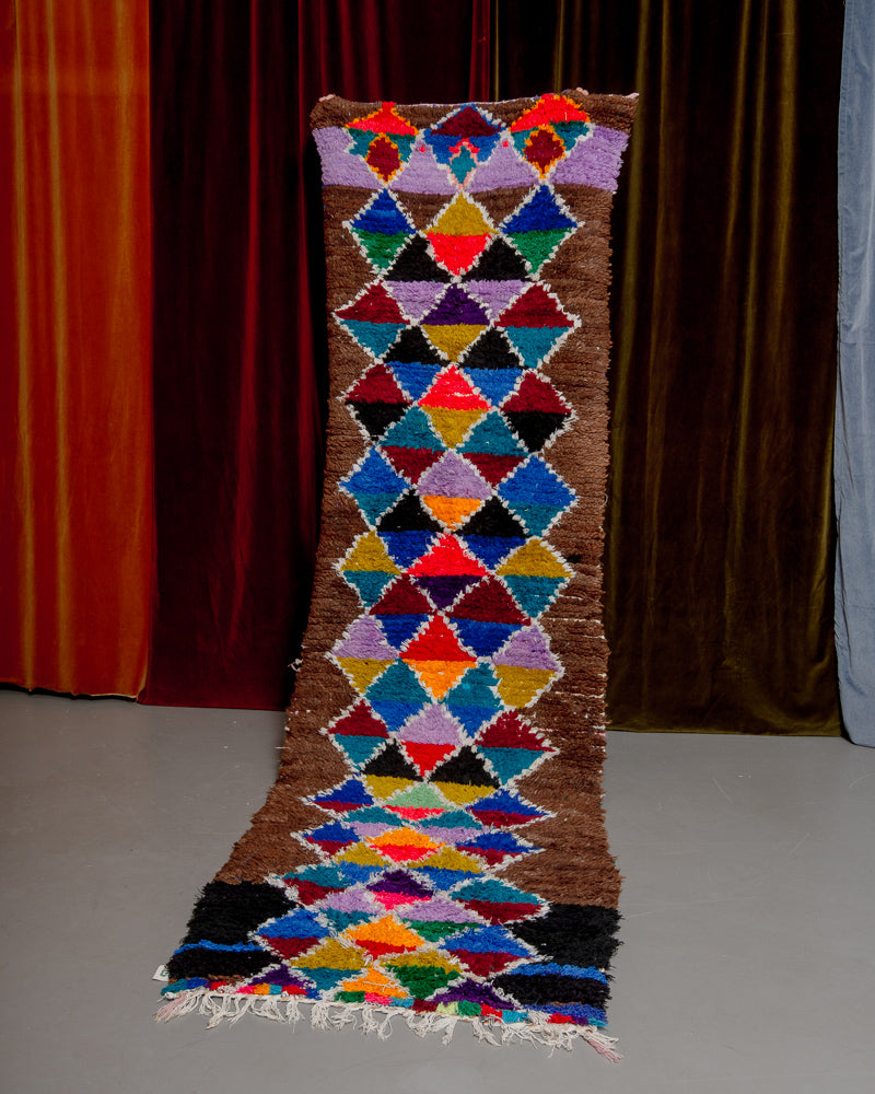 Vintage Berberlin boucherouite runner rug being held up in front of a velvet curtain. 
