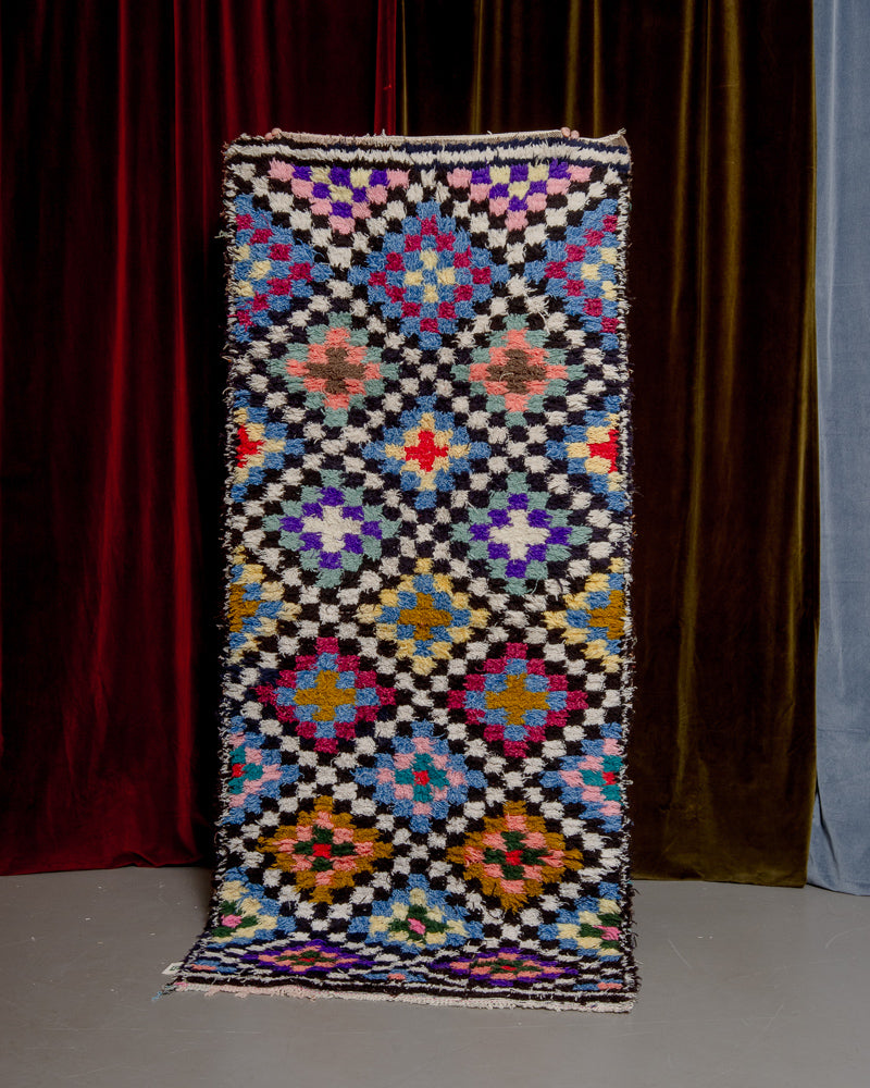 Colourful boucherouite vintage rug with a diamond pattern made up of small squares being held in front of velvet curtains.