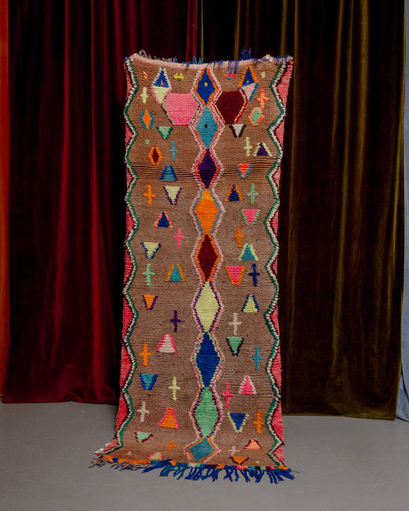 Vintage Berberlin boucherouite runner rug being held up in front of a velvet curtain. 