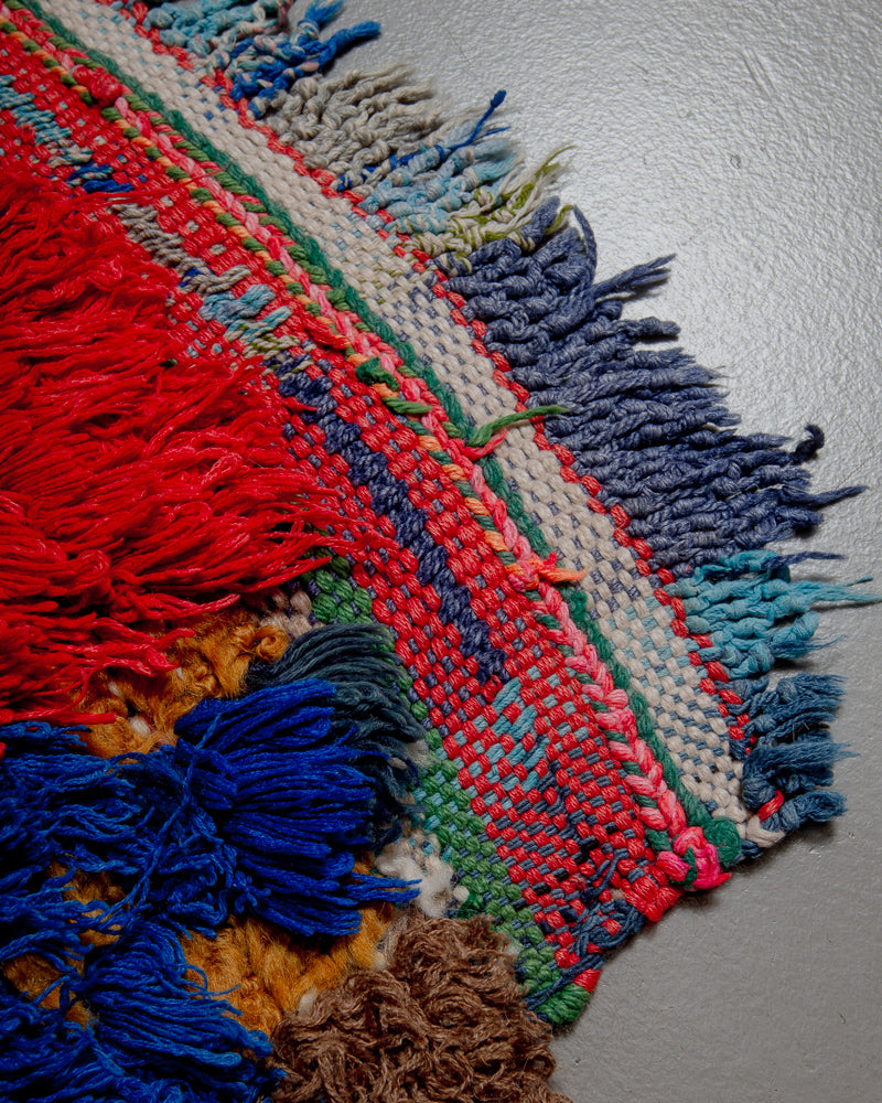 Corner of boucherouite rug with red, blue green and white yarn and tassels.