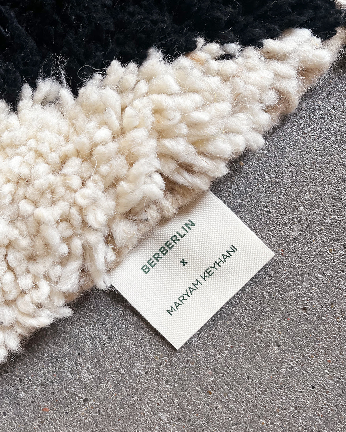 Cotton label with Berberlin x Maryam Keyhani printed on it in green, sewn onto the edge of a cream and black wool Beniourain rug. 